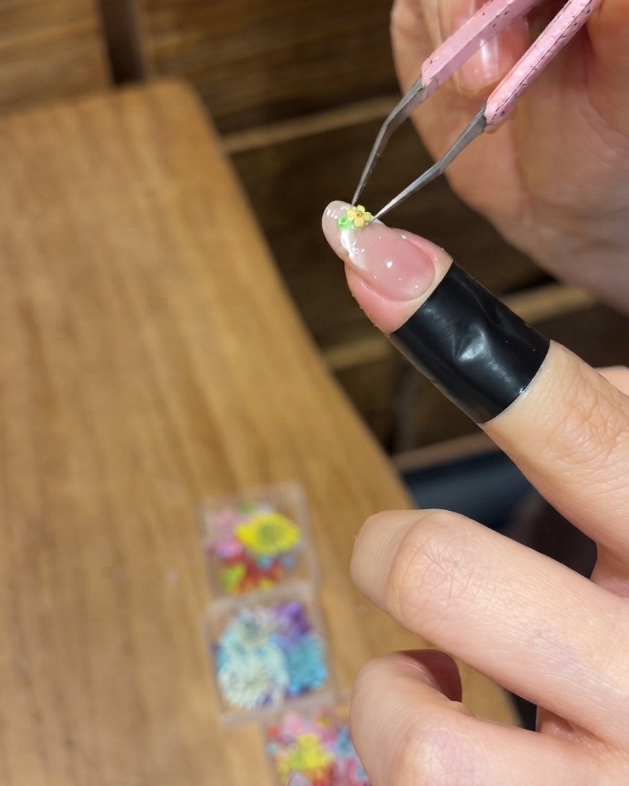 Pinzas de precisión colocando una pequeña flor seca amarilla sobre la superficie húmeda de gel de una uña.
