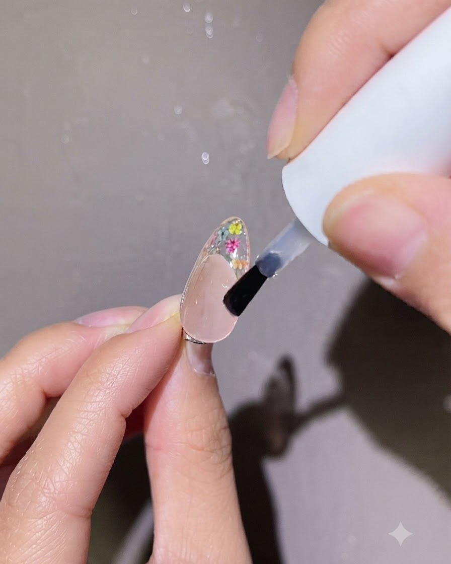 Un pincel pequeño aplicando un punto preciso de capa superior transparente sobre el arte de uñas floral en una uña.