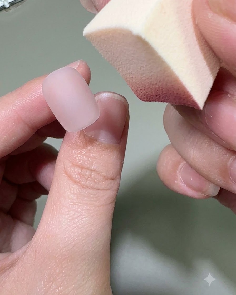 Mano sosteniendo una esponja de maquillaje aplicando color de gel rosa en la punta de una uña nude mate.
