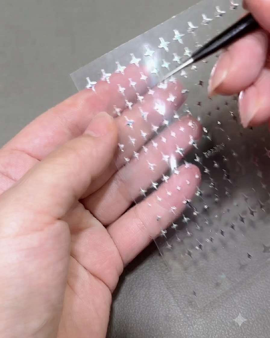 Primer plano de una herramienta de detalle levantando una diminuta pegatina de estrella plateada de una hoja transparente sostenida sobre la palma de una persona.