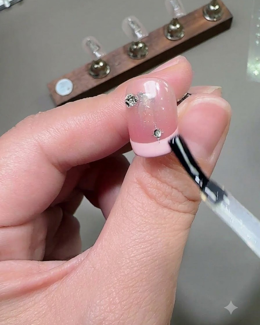 Un pincel aplicando una capa de top coat brillante sobre una uña decorada con pedrería y dijes de estrellas.