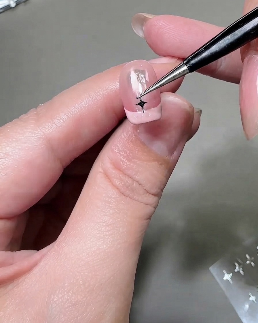 Una herramienta fina posicionando una pegatina de estrella plateada en la punta de una segunda uña con manicura francesa.