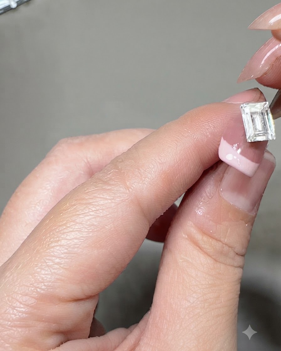 Un par de pinzas colocando una gema de cristal rectangular grande en el centro de una uña con manicura francesa.