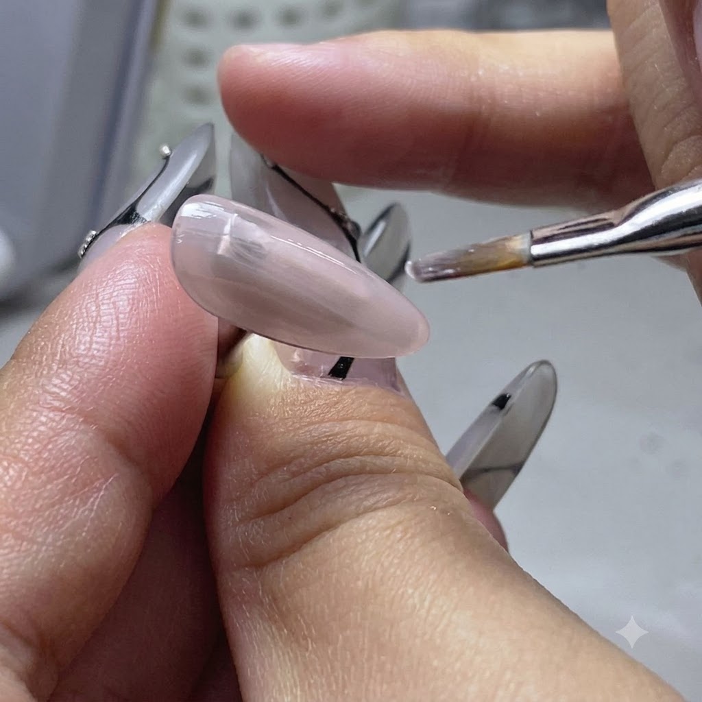 Un pincel plano aplicando una capa fina de gel base de color nude sobre un dedo de práctica para uñas.