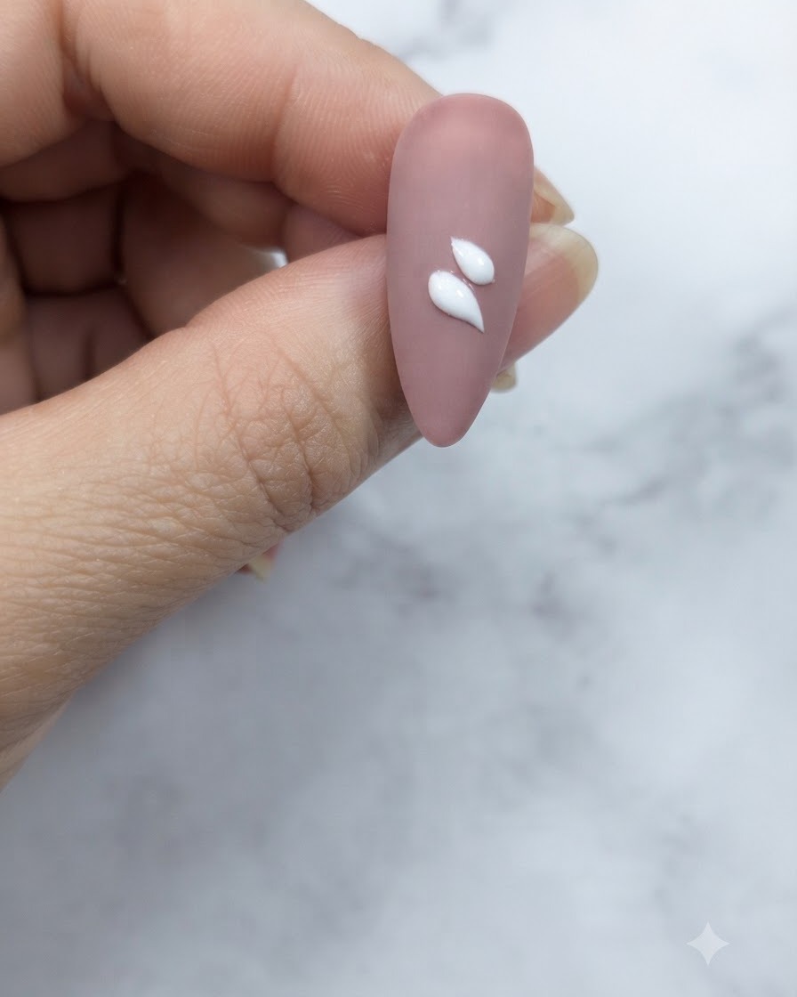 Un primer plano de una uña de color nude rosado que muestra dos trazos de pétalos blancos distintos en forma de lágrima aplicados cerca del centro.