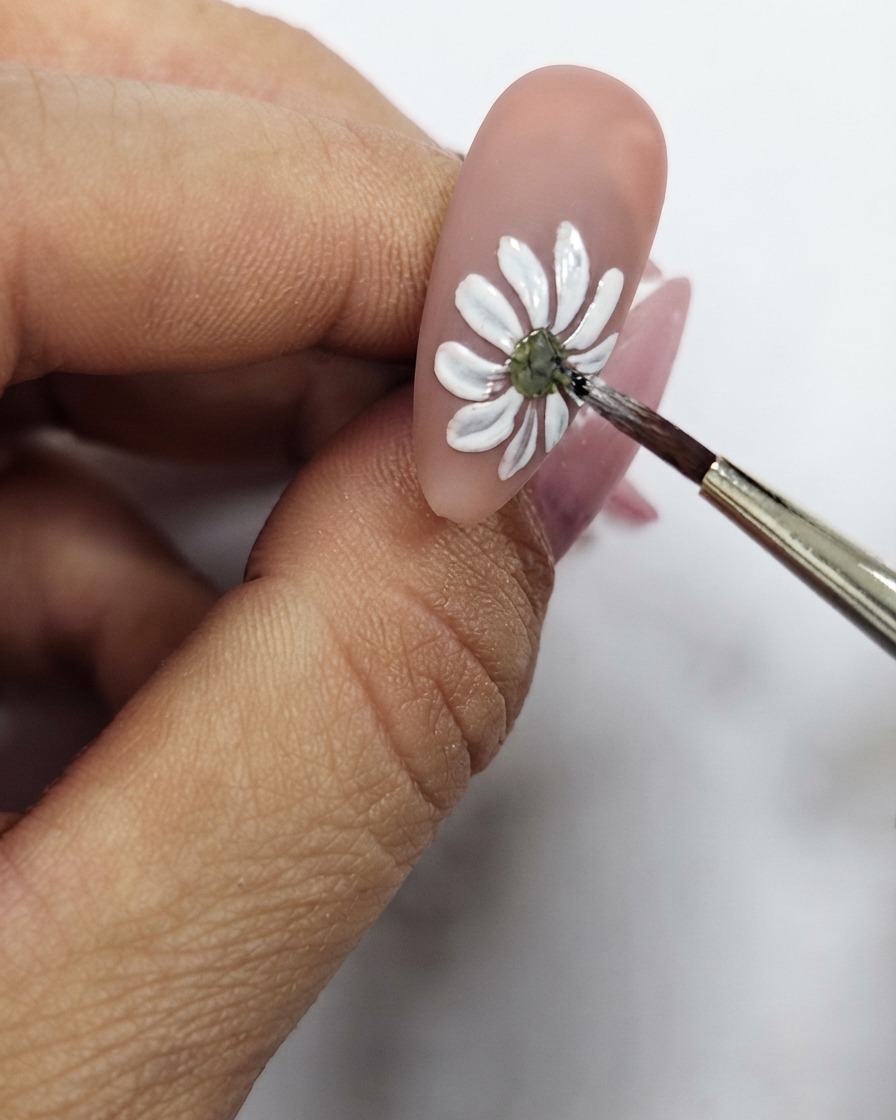 Un primer plano de un pincel fino refinando el punto central amarillo del diseño de uñas de margarita blanca.