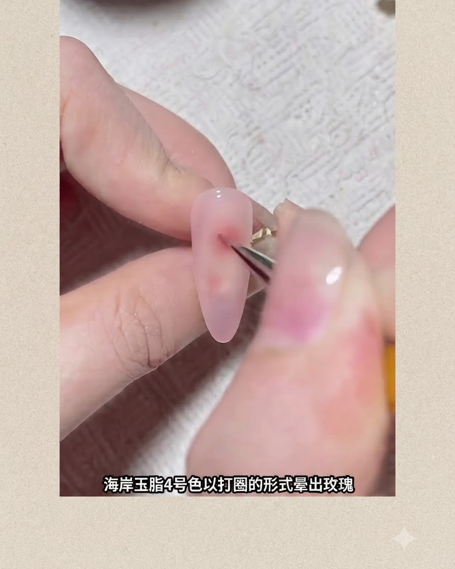 Un pincel fino de arte de uñas dibuja un punto de pigmento circular rojizo sobre una uña recubierta de gel rosa para formar una flor en expansión.