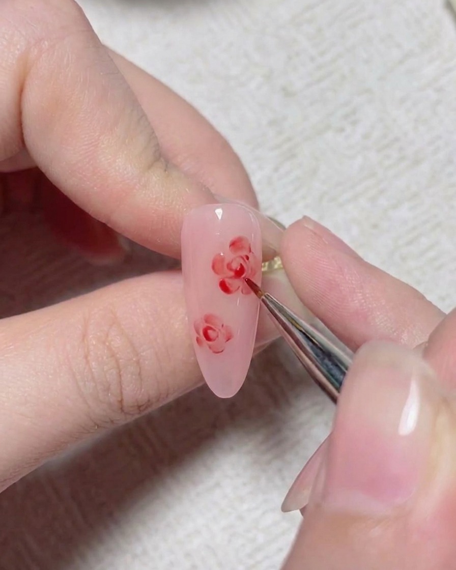 Un pincel fino de arte de uñas agrega líneas de detalle rojas para formar el centro de una rosa pintada sobre una uña rosa.