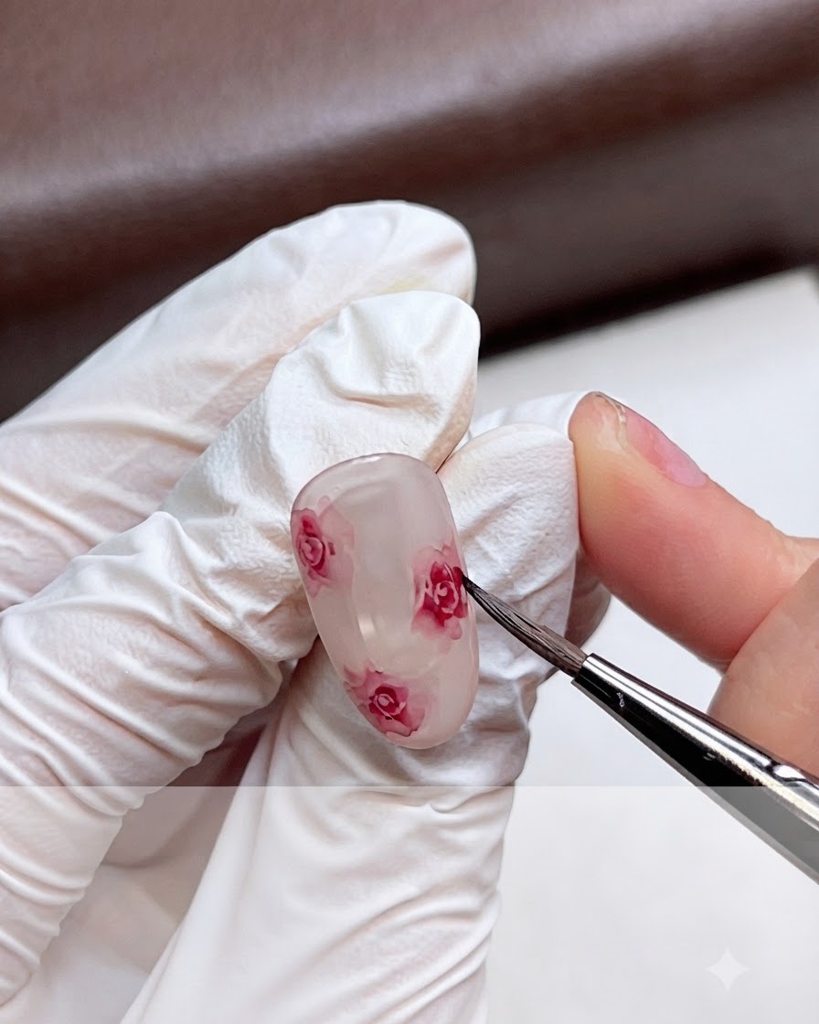 Una mano con guantes difuminando suavemente los bordes de una rosa de acuarela rosada en una uña de gel usando un pincel fino.