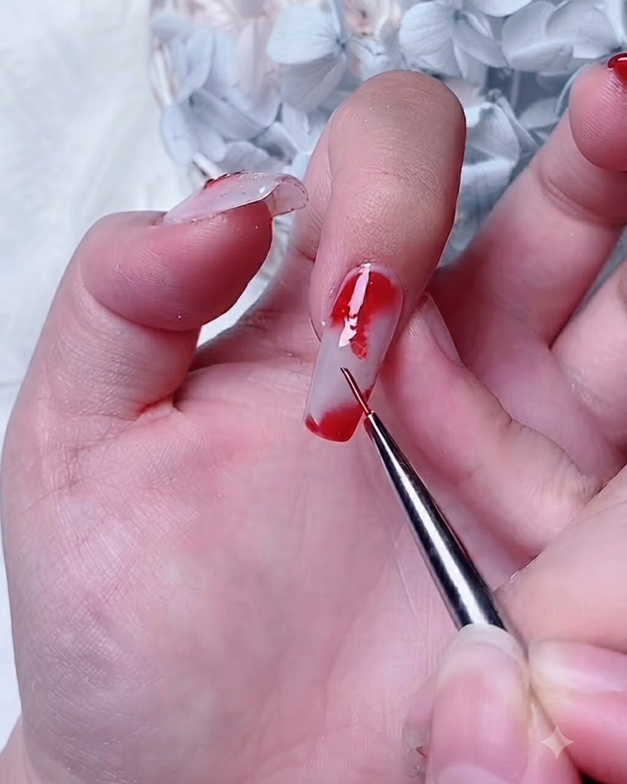 Un pincel de detalle aplicando gel rojo sobre una capa de gel blooming en la esquina de la uña del índice.
