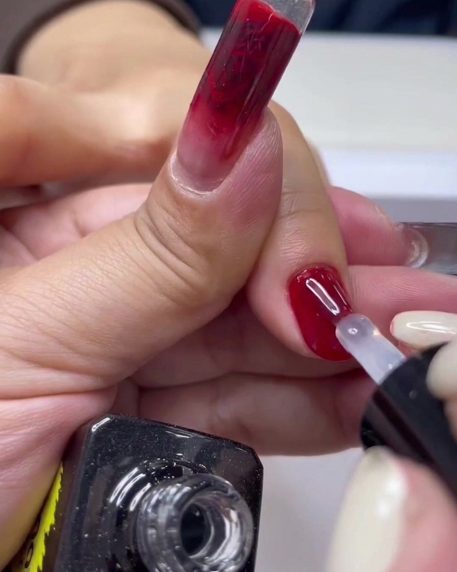Un primer plano de un pincel de uñas aplicando top coat transparente sobre una manicura de gel roja brillante.