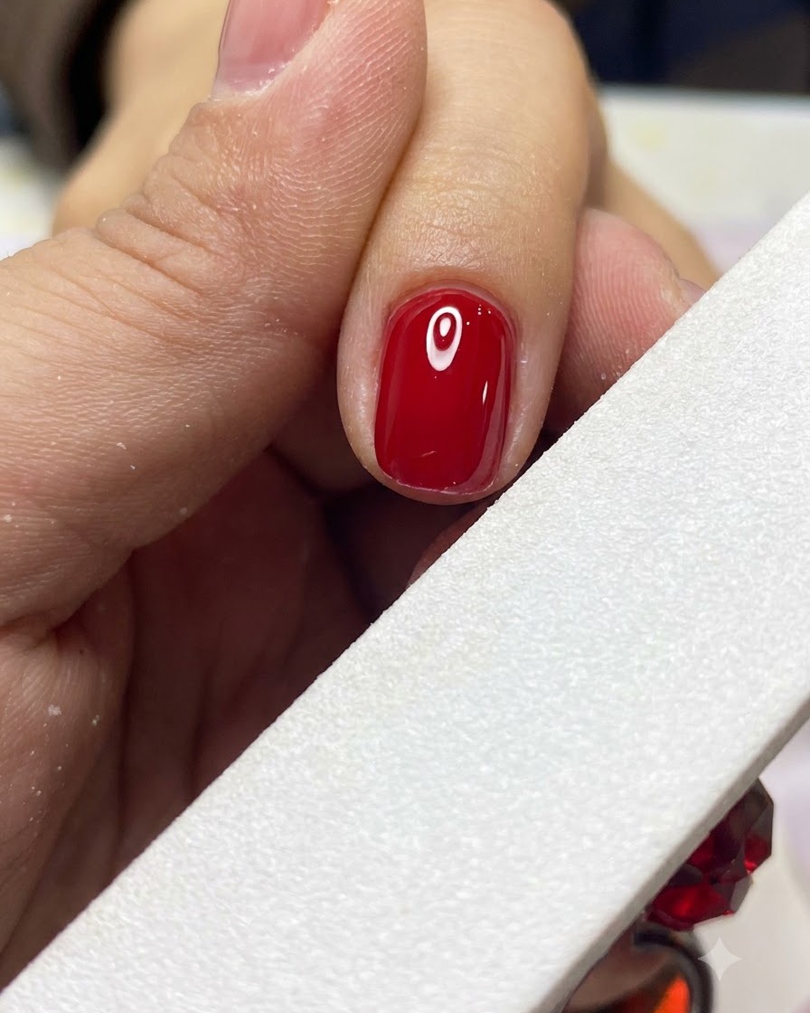 Una lima de uñas blanca dando forma y suavizando suavemente el borde de una uña de gel roja terminada.