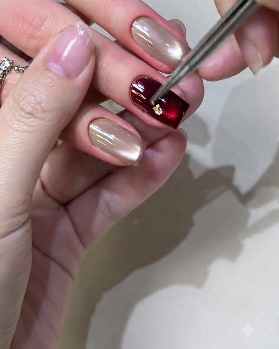 Unas pinzas colocando pequeños trozos de lámina de oro sobre una uña con esmalte de gel borgoña.