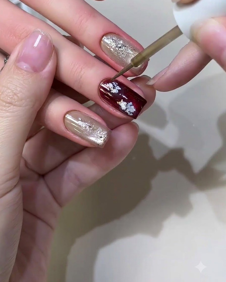 Un pincel fino de nail art aplicando líquido metálico para formar pequeños patrones florales casuales en una uña borgoña.