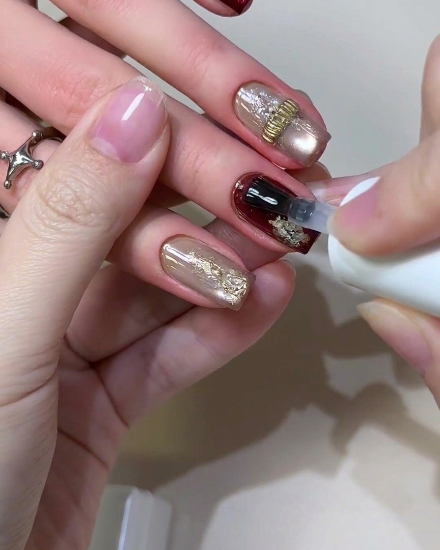 Un pincel de gel aplicando una capa superior selladora transparente y brillante sobre un diseño completado de uñas retro en borgoña y lámina dorada.
