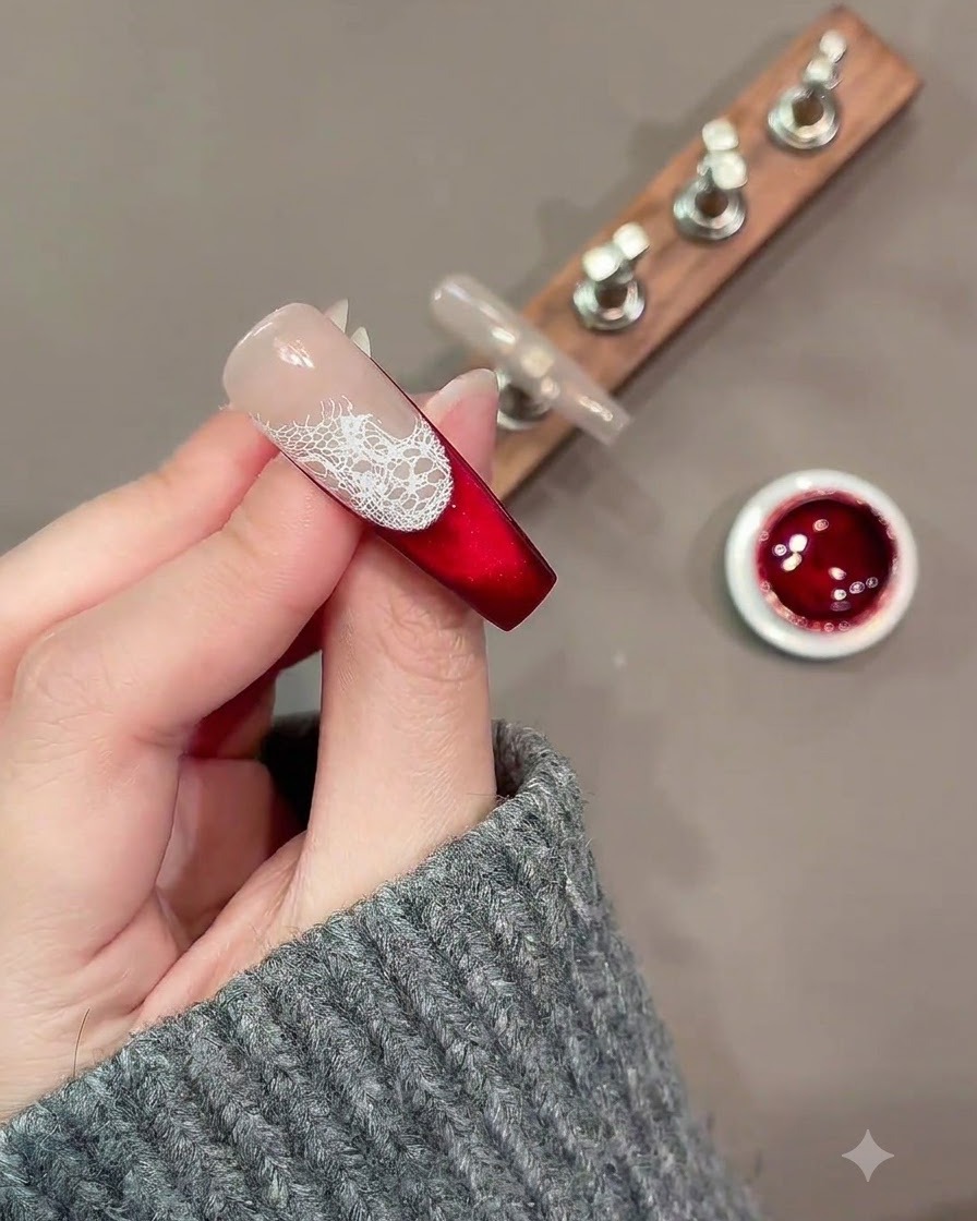 Un pincel fino de arte en uñas pintando contornos de rosas blancas sueltas sobre un patrón de encaje en una uña roja y nude.