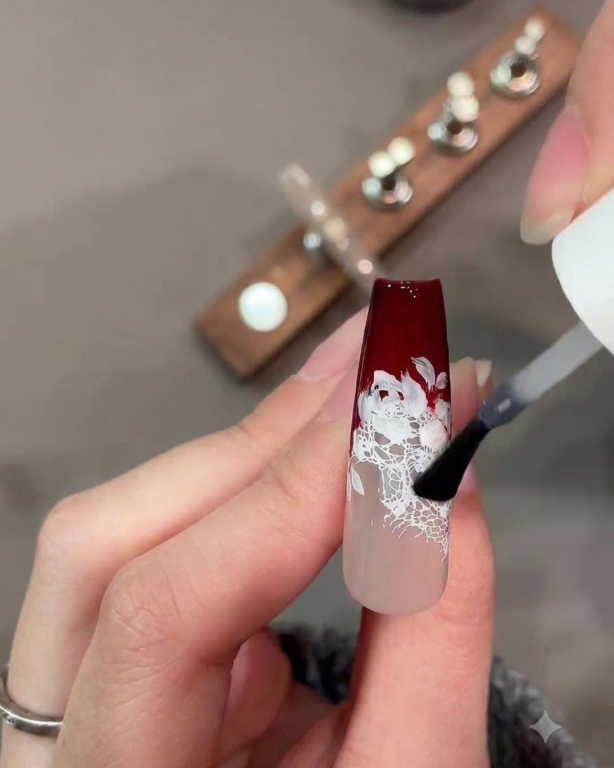 Un pincel aplicando una capa superior mate sobre un diseño de uñas de encaje rojo oscuro y blanco.