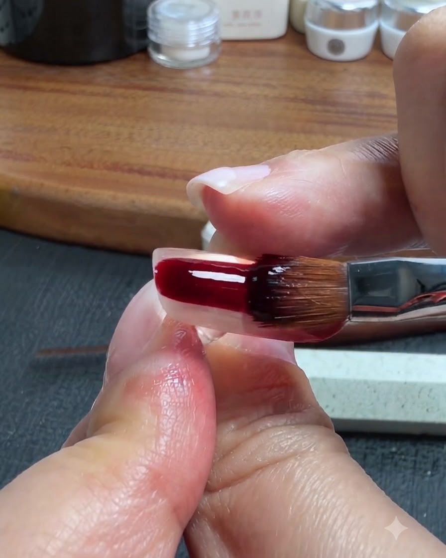 Un pincel de uñas aplicando un gel translúcido rojo oscuro y marrón sobre la porción inferior de una uña artificial transparente.