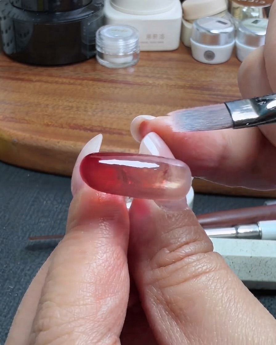 Un pincel de maquillaje limpio difuminando suavemente el borde de una capa de gel translúcido rojo en una uña.