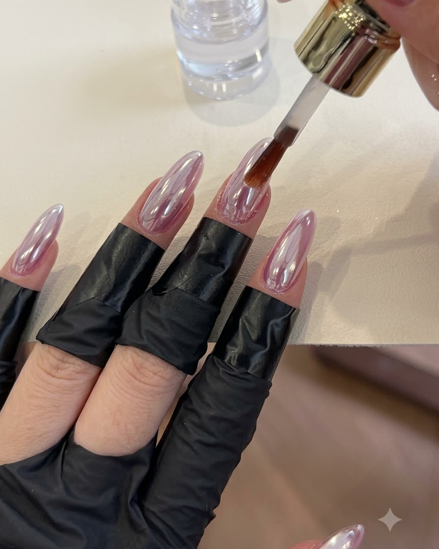 Un pincel aplicando una capa superior transparente sobre uñas brillantes con acabado cromado.