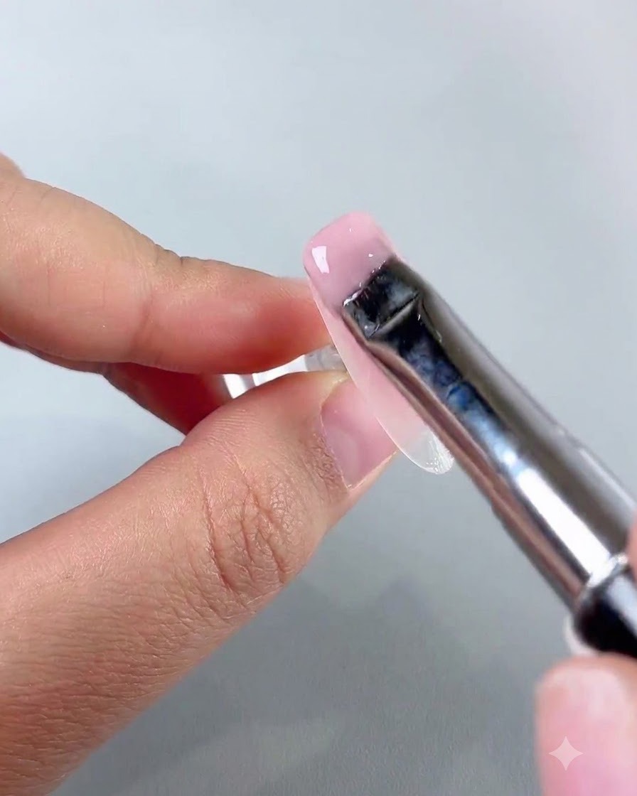 Un pincel plano de nail art esparciendo una capa suave de gel base jade rosa claro sobre la superficie de la uña.
