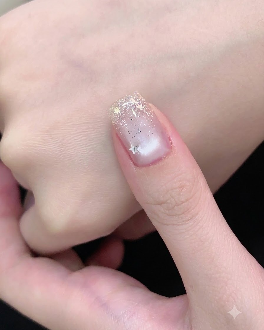 Un pincel de uñas aplicando una capa superior transparente sobre una uña decorada con brillo plateado y piezas de estrella.