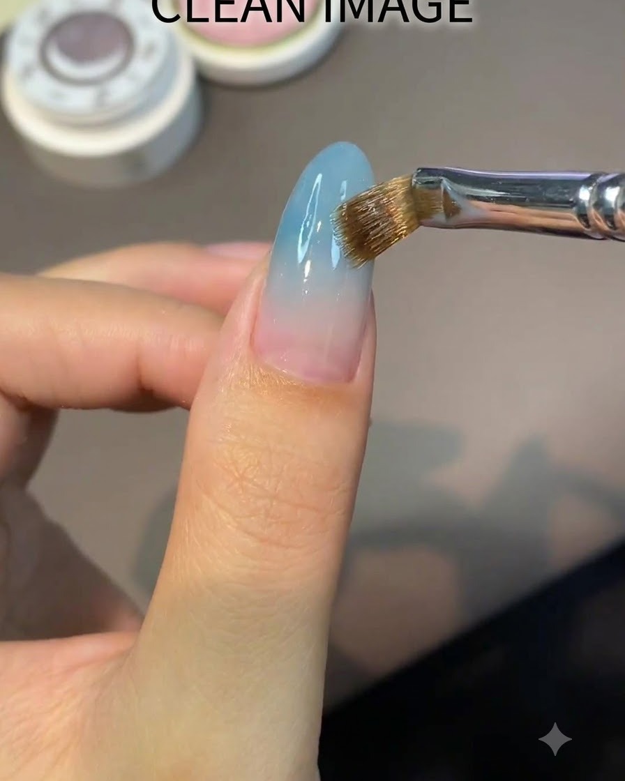 Un pincel plano aplicando una capa uniforme de gel base transparente nude sobre una uña en forma de almendra.