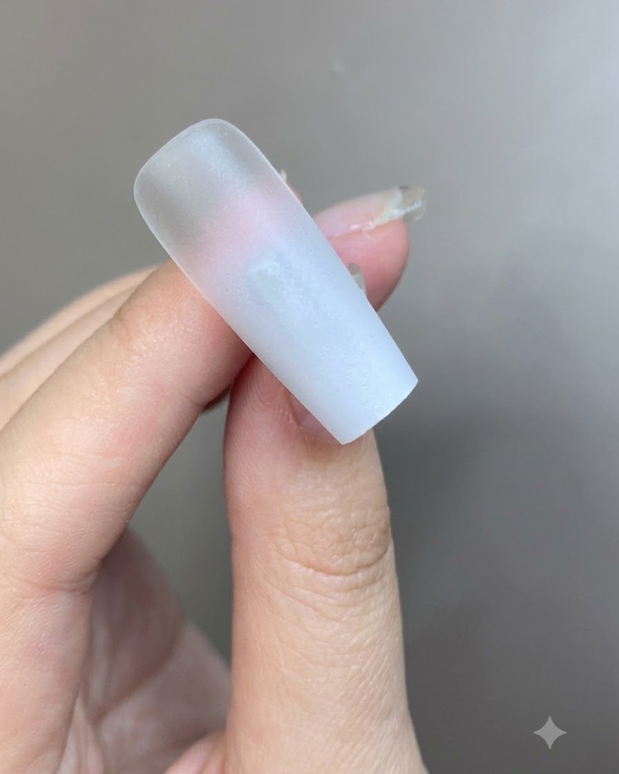 Una capa fina de gel blanco lechoso siendo aplicada con pincel sobre la superficie de una uña degradada.
