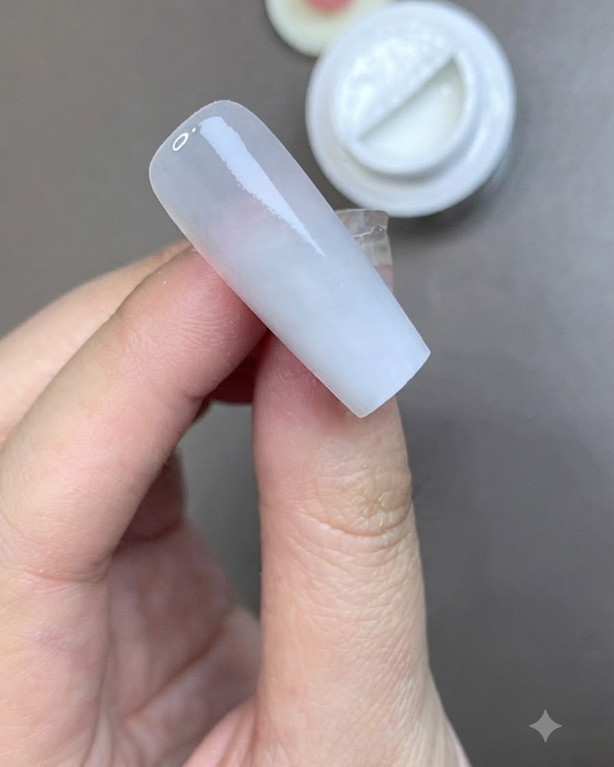 Un pincel aplicando una capa suave de brillo superior mate sobre una uña preparada en color blanco lechoso.