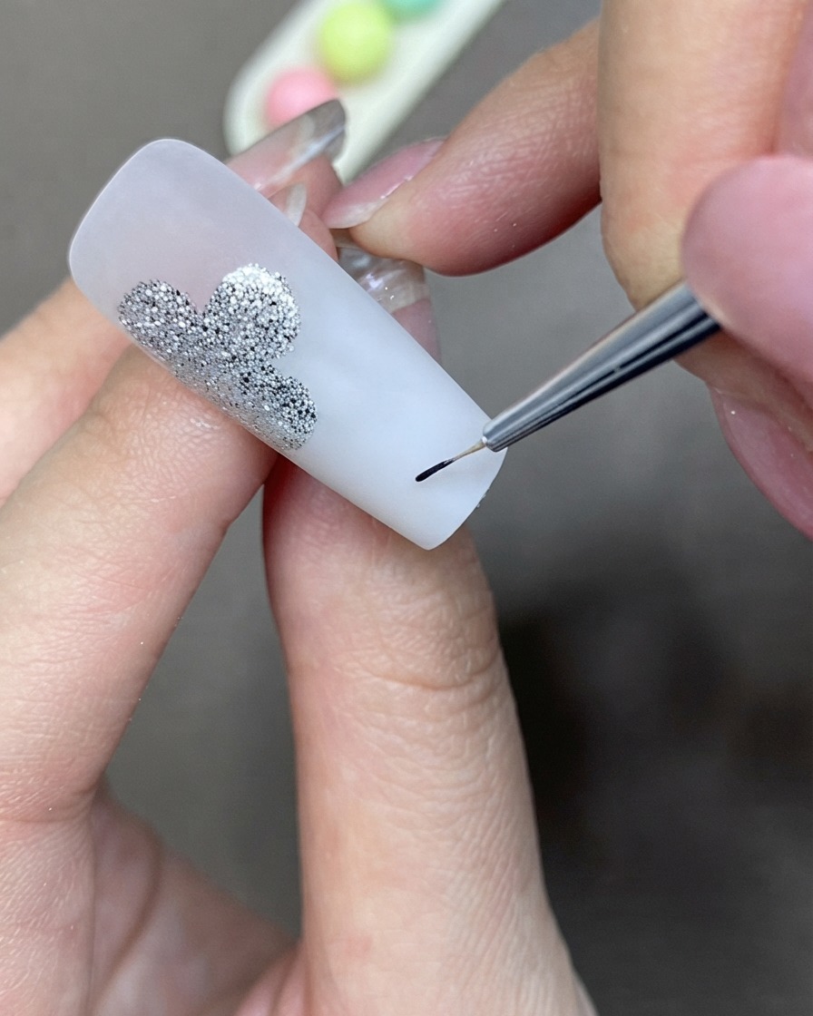 Un pincel fino de detalle aplicando gel delineador negro para crear una forma de trébol junto a un diseño de purpurina plateada.