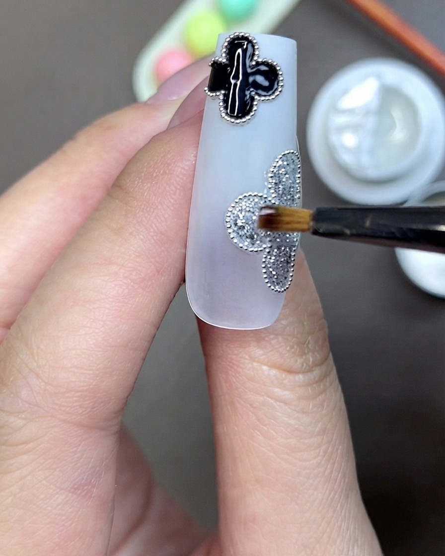 Un pincel aplicando gel adhesivo transparente para pedrería en el centro de un motivo de trébol de purpurina plateada en una uña.