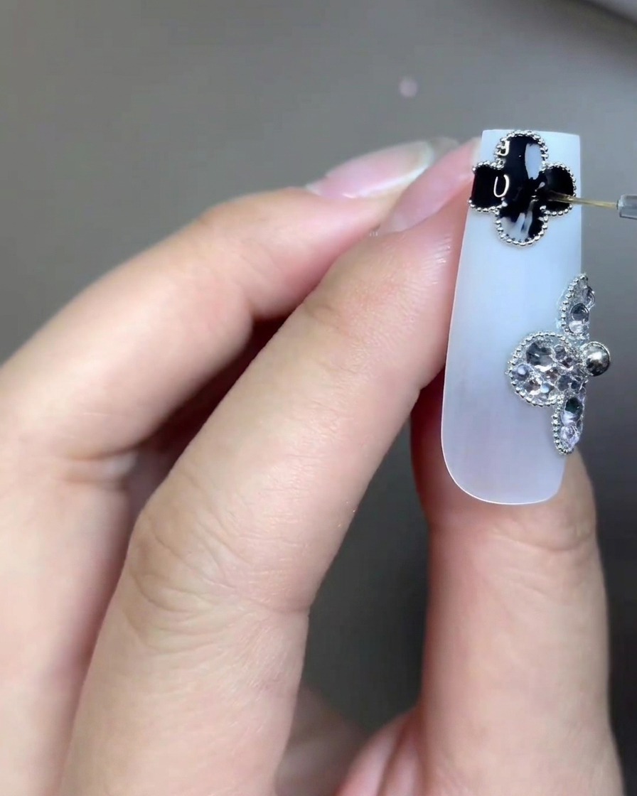 Un primer plano de una uña larga de color blanco lechoso adornada con dos dijes de trébol de cuatro hojas: uno negro, otro hecho de pedrería. Un pincel de arte de uñas muy fino aplica una capa superior transparente con precisión sobre el detalle del trébol negro.