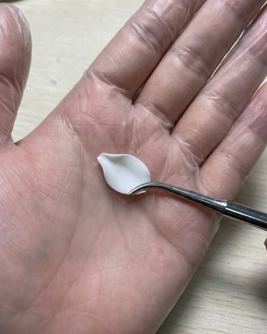 Una mano enguantada sosteniendo una herramienta de silicona alisando un pétalo de gel 3D blanco que descansa sobre un tapete de silicona.