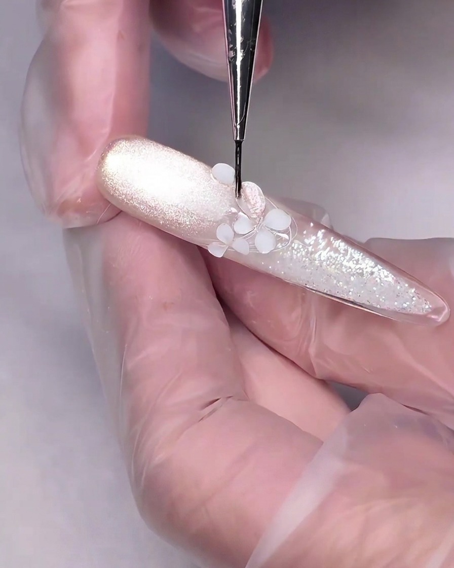 Un pincel de detalle fino aplicando gel ojo de gato rosa nude sobre una flor esculpida en 3D blanca en una uña.