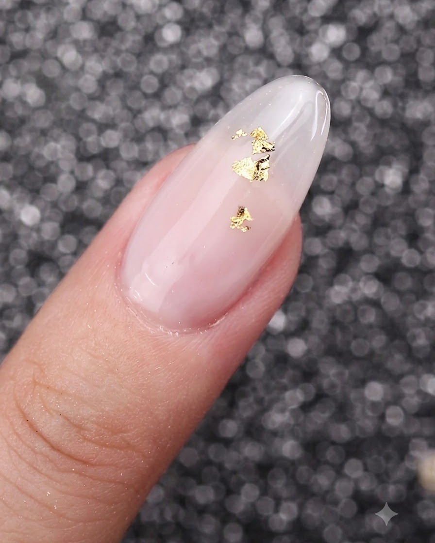 Una vista de cerca de copos de lámina dorada colocados delicadamente sobre una uña con manicura de gel blanco lechoso.