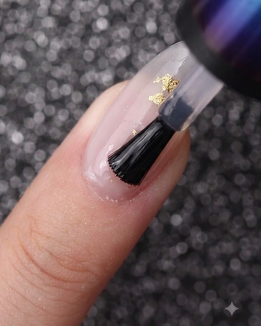Un pincel de uñas extendiendo gel transparente sobre una uña decorada con esmalte blanco lechoso y lámina dorada.