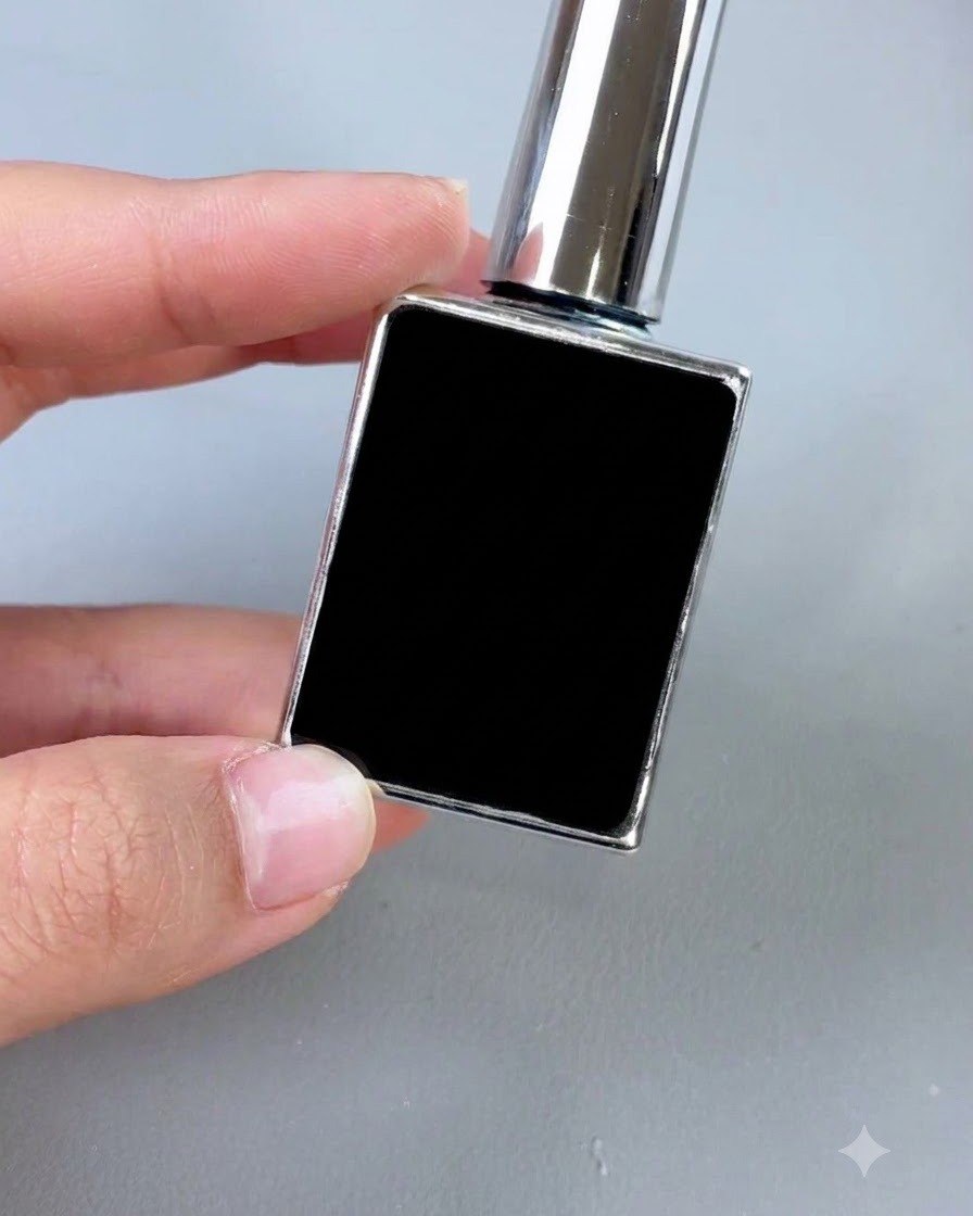 Un frasco cosmético negro pequeño con una tapa plateada y un pincel aplicador, mostrado en posición vertical contra un fondo gris.