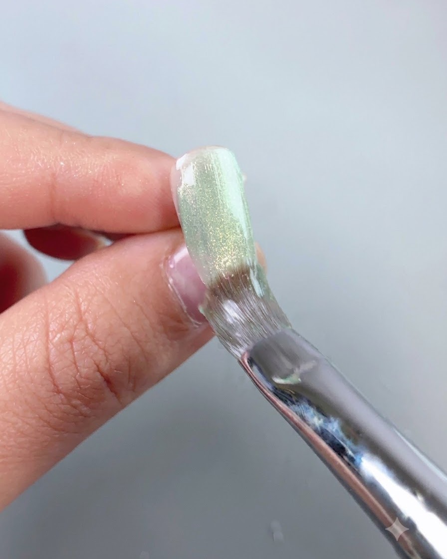 Un pincel de uñas que suaviza una capa de gel ojo de gato verde transparente sobre la superficie de una uña postiza.