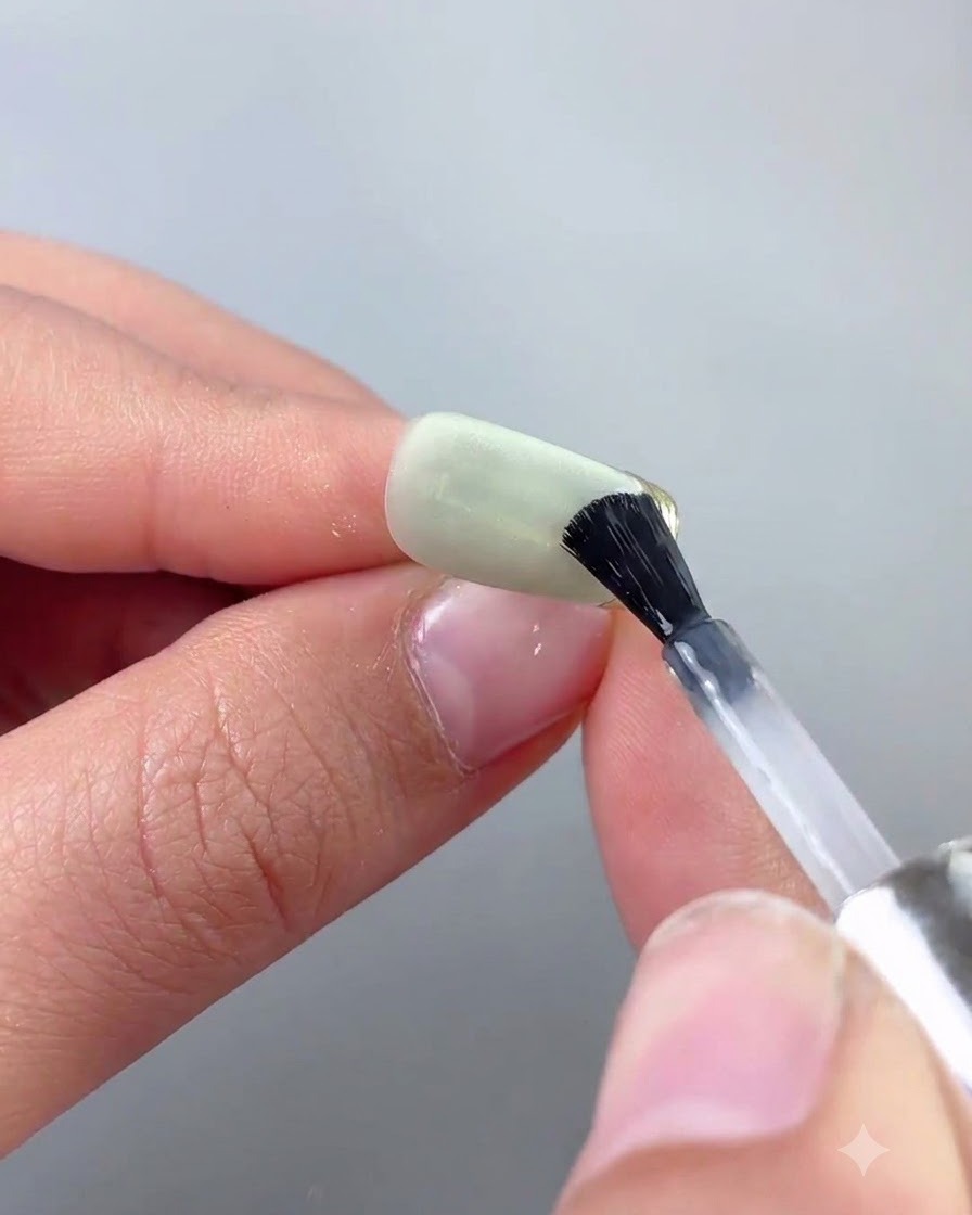 Un pincel fino de arte de uñas que aplica una capa superior precisamente a lo largo de un borde de cromo dorado de una uña de ojo de gato verde.