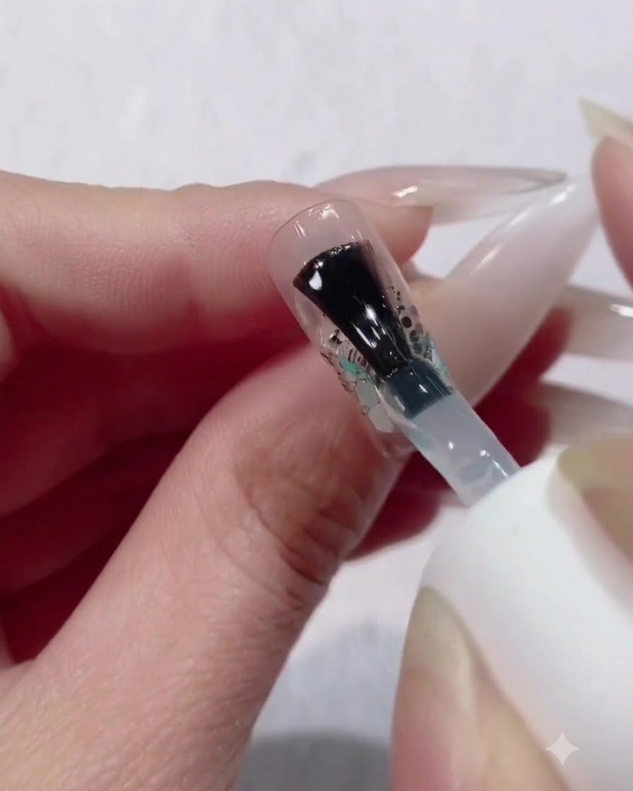 Un pincel aplicando una capa gruesa y generosa de gel constructor transparente sobre un diseño de uñas con degradado de purpurina y lentejuelas.