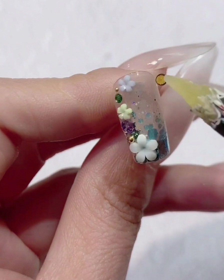 Un primer plano de una mano sosteniendo una uña decorada con gel transparente, purpurina y pequeñas flores 3D, mientras una herramienta de cera coloca una pequeña pedrería amarilla en el diseño.
