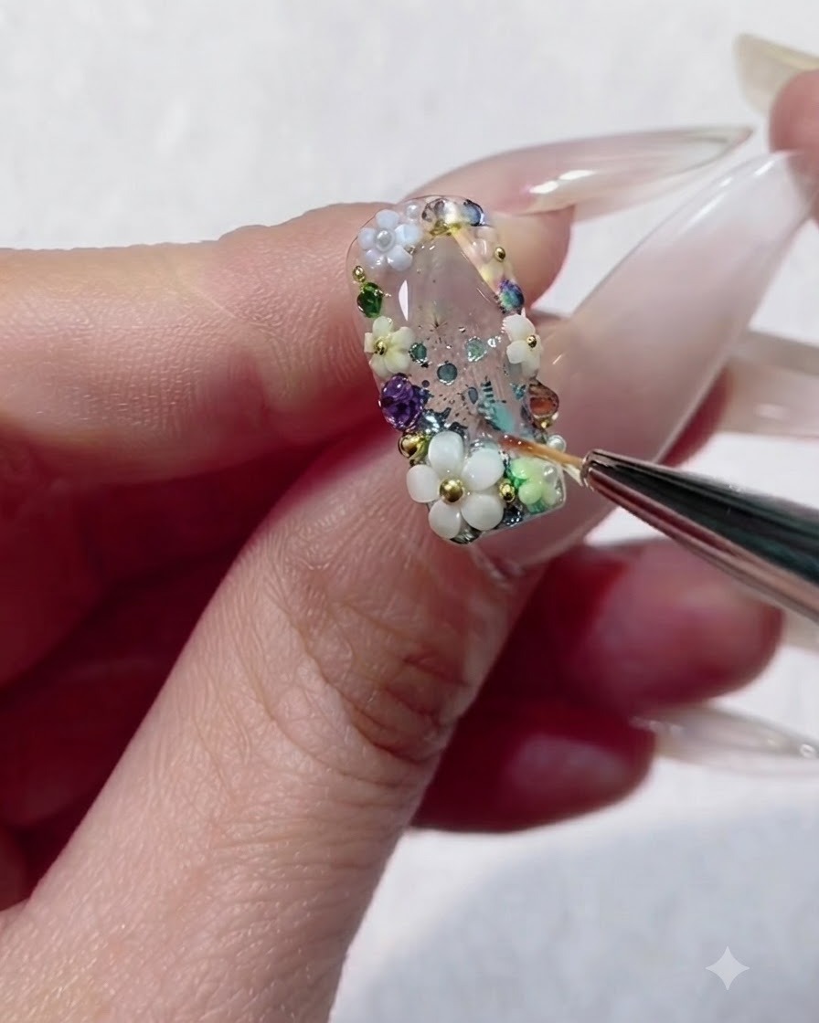 Un pincel delineador inundando los diminutos espacios entre pedrería colorida y cuentas en una uña de gel transparente.