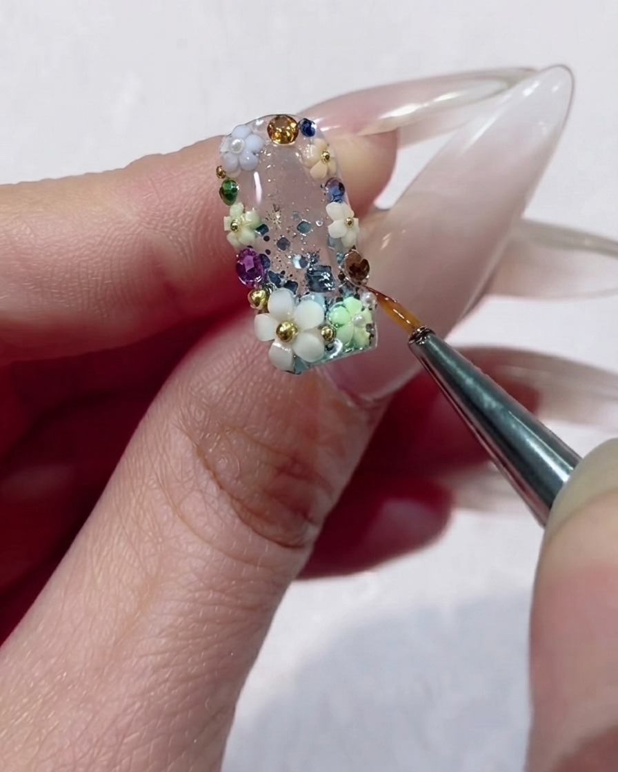 Un pincel delineador fino aplicando con precisión gel transparente alrededor de los bordes de una pedrería facetada en un diseño de uñas floral.