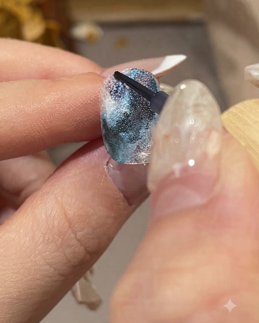Primer plano detallado de una mano manicurada sosteniendo un pincel fino para aplicar gotas de tinta azul oscura sobre una superficie de uña texturizada.