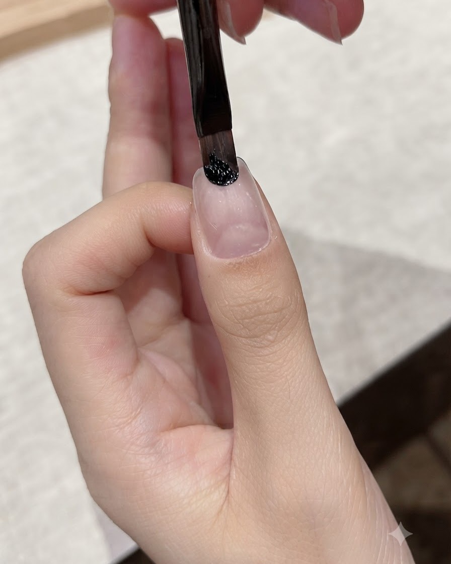 Un pincel delineador fino aplicando gel negro oscuro cerca de la zona de la cutícula de una uña natural.