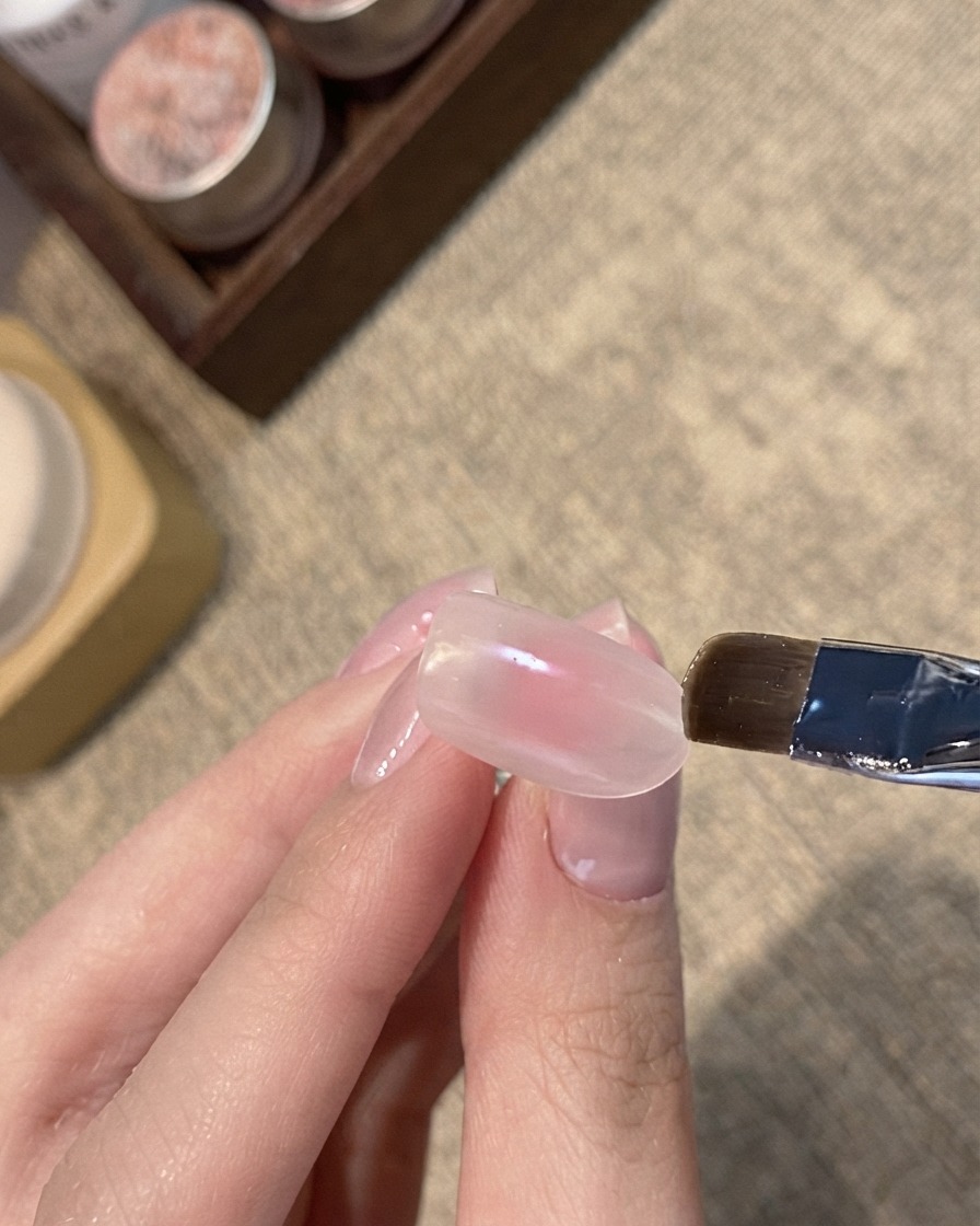 Un pincel plano aplicando gel cat-eye con brillo azul sobre la punta de una uña con tono nude rosado.