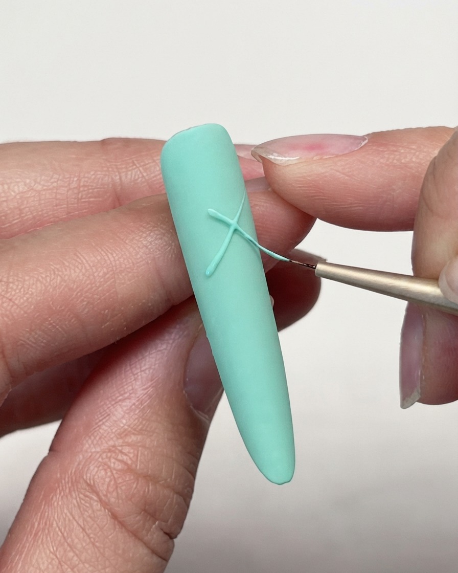 Un primer plano de un pincel delineador fino cruzando líneas de gel en un tip de uña mate verde menta para comenzar un diseño de estrella.