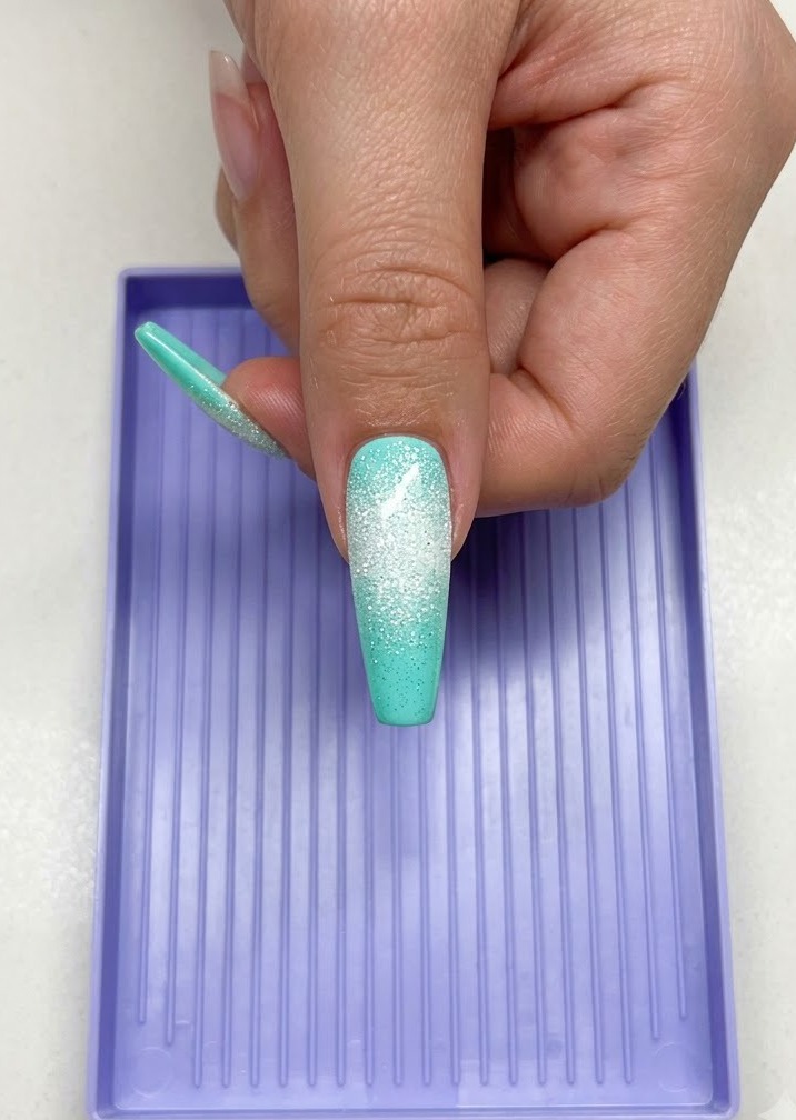 Una mano sosteniendo una uña larga de color menta con un diseño de estrella azucarada blanca debajo de una lámpara de curado UV LED morada.