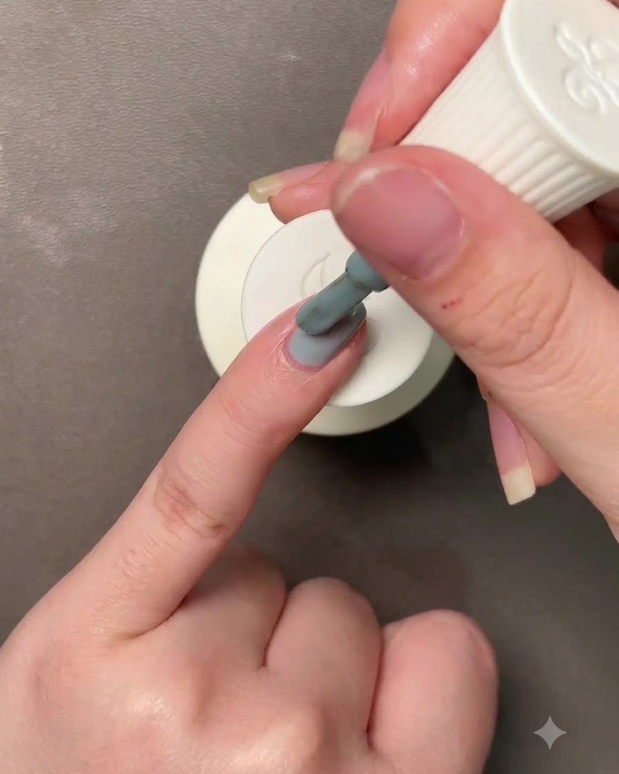 Una mano sosteniendo un pincel aplicando un esmalte de gel azul verdoso Morandi grisáceo a una uña.