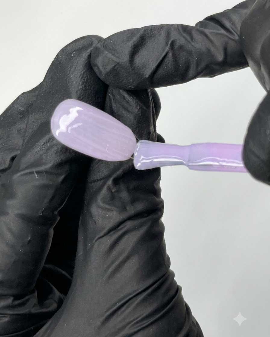 Un pincel plano deslizando el esmalte de gel morado traslúcido N04 de manera uniforme sobre la superficie de una punta de uña de práctica ovalada.